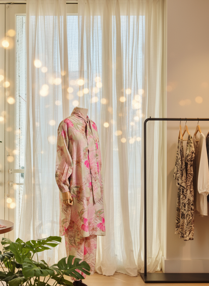 Mannequin wearing a floral dress in a room with white curtains and a plant.