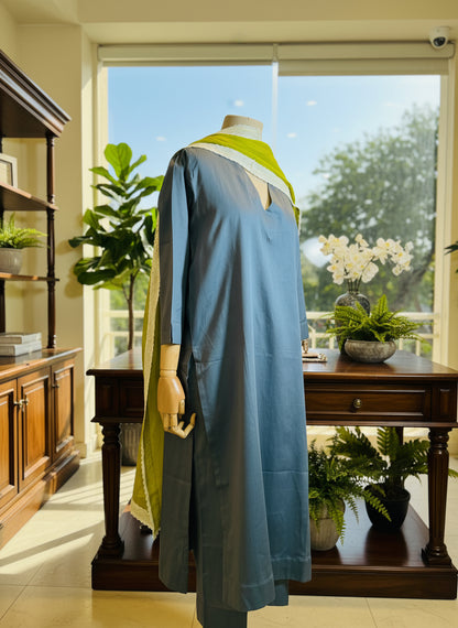 Mannequin wearing a blue outfit with a green sash in a room with curtains.